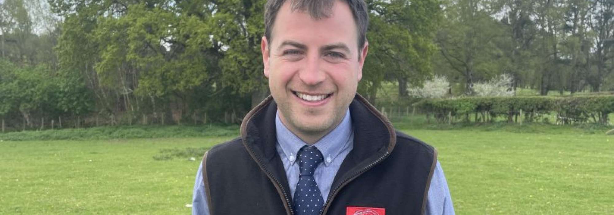 Man with dark hair standing outside in a shirt and gilet in front of a tree.