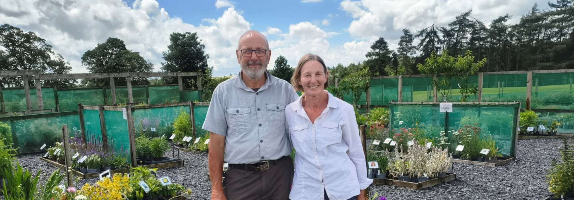 Two people standing outside surrounded by flower beds and gravel. 